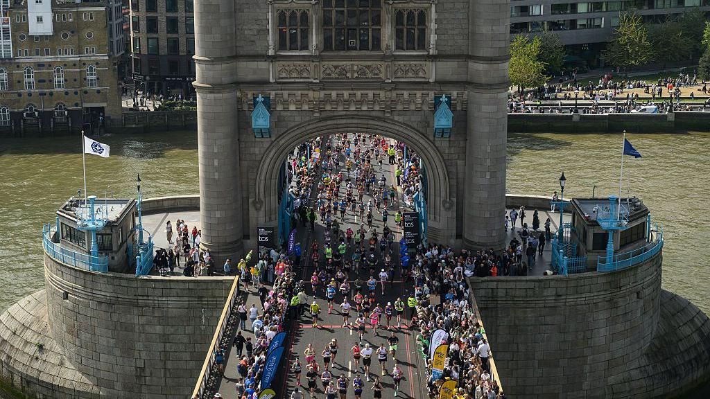 An aerial view of a sea of marathon participants running across Tower Bridge, with the River Thames visible on either side.