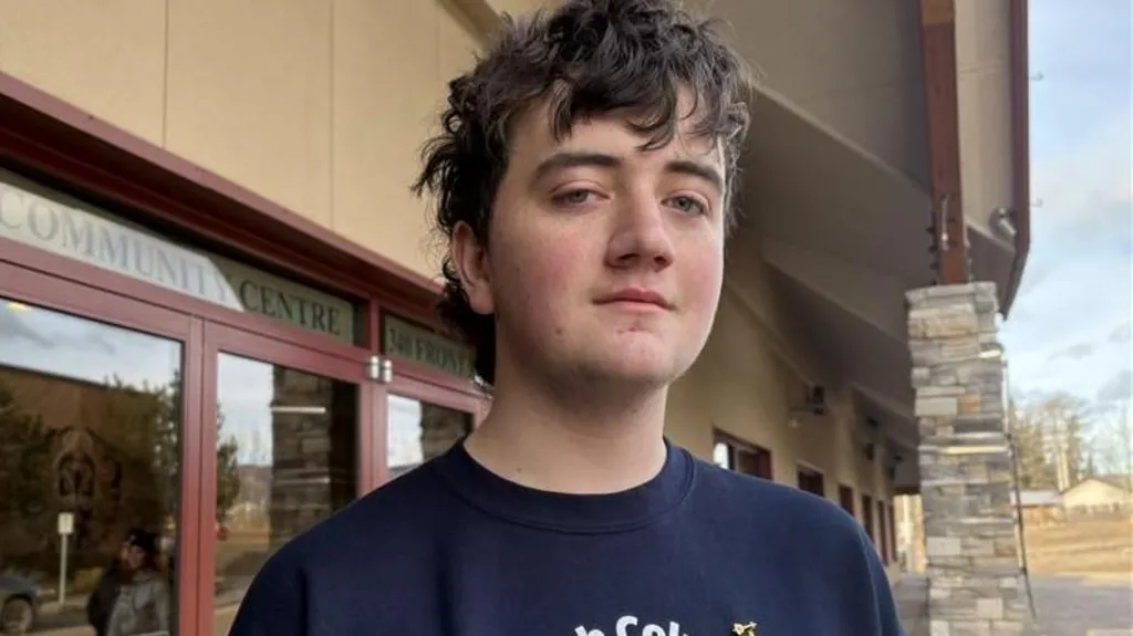 Darian Quist in a navy blue T-shirt looks at the camera with a neutral expression. Behind him is a community centre.  