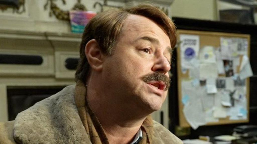 Danny Dyer's moustached character looks off camera and has a some-what pleading look on his face and his arms outstretched. The scene behind him is a messy office with lots of files, desks, computers and blue lamps. He's wearing thick gloves and a warm lambskin jacket.