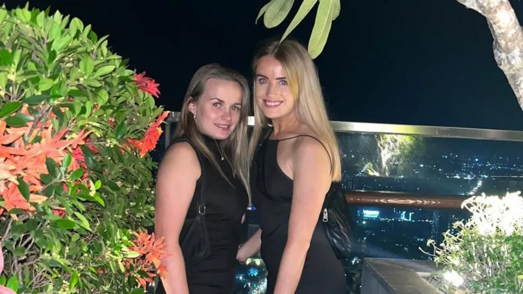 Two women, wearing all-black dresses, posing on of a balcony at night, surrounded by some plants. They're both smiling at the camera.  