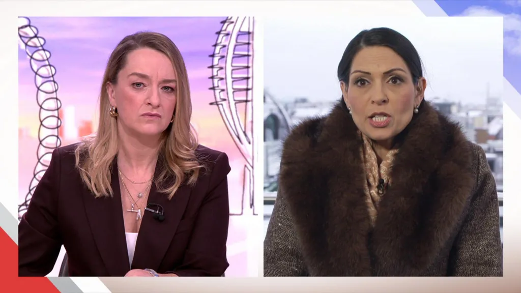 Laura Kuenssberg interviews Dame Priti Patel, who is at the Munich Security Conference. Kuenssberg is in the BBC studio, wearing a dark maroon suit and a white top, with multiple necklaces and her long, blonde hair worn loose. Patel is standing on a balcony with snow-covered buildings behind her. She wears a brown coat with a large, chocolate brown fur collar and a light brown scarf underneath. She wears her dark hair pulled back into a low ponytail with two pearl earrings. 