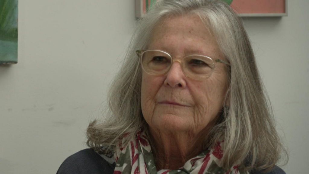 Susanna Rook wears glasses, a dark coloured top and a patterned scarf around her neck. She is seated and there is some artwork behind her.