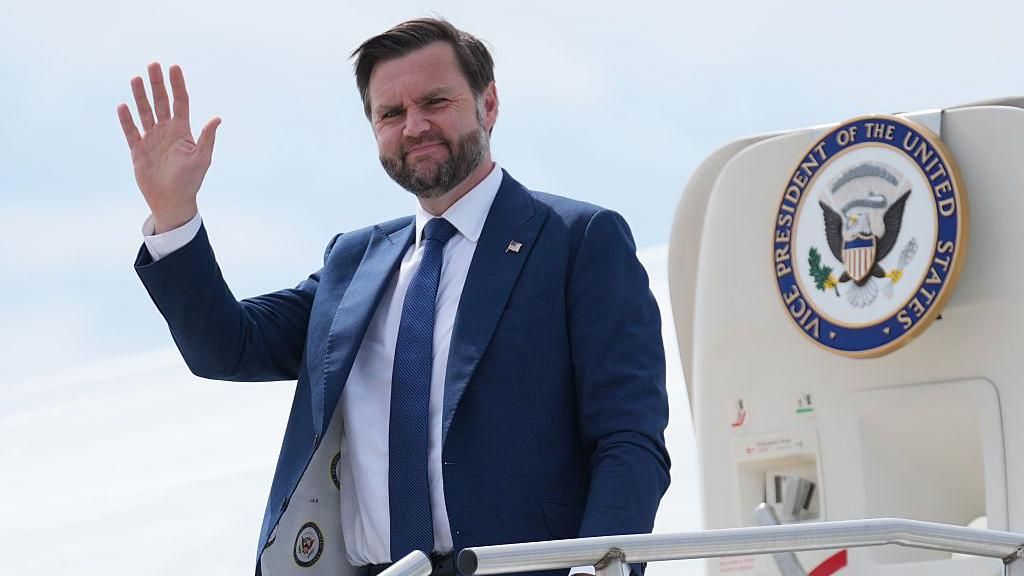 JD Vance waving as he steps out of a jet with the presidential seal on the door. He wears a blue suit with a white shirt and blue tie, and has a pin on his lapel with the US flag on it.