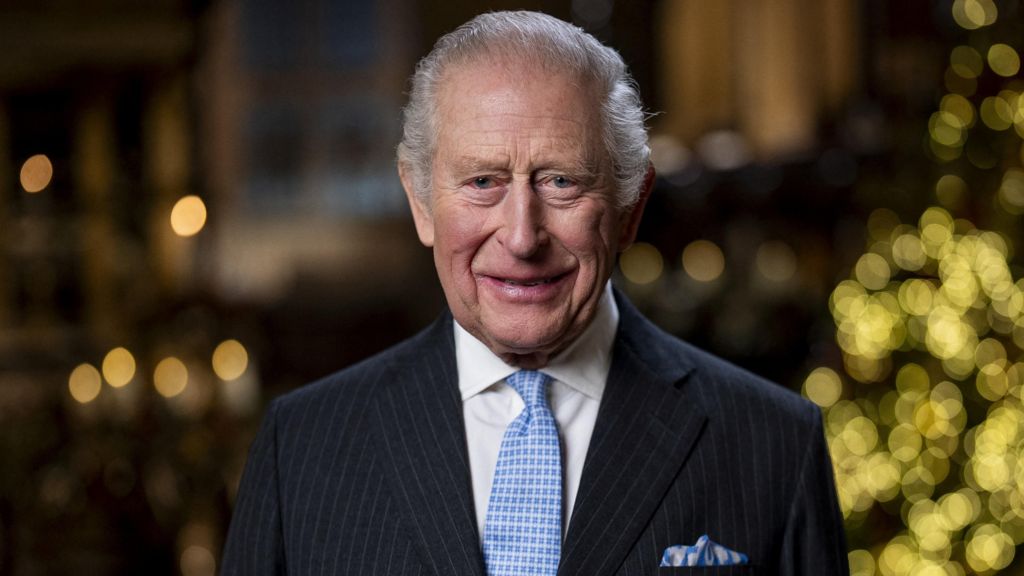 King Charles, wearing a pinstriped suit and pale blue tie and pocket square, smiles at the camera, with the lights from a Christmas tree just seen in the background