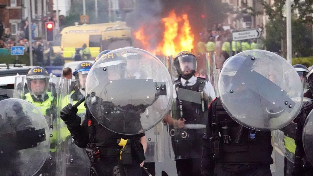 Police officers wearing riot gear, facing the camera with shields and batons raised, while in the background a police van in burning