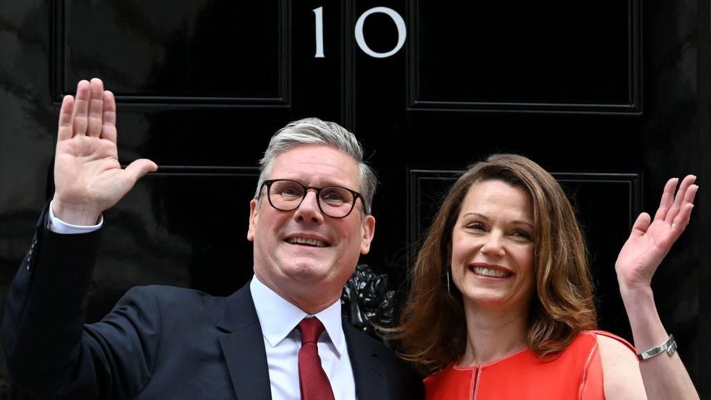 Sir Keir Starmer, in a navy blazer, red tie and white shirt, and his wife Victoria, in a red dress, wave outside the door of 10  Downing Street