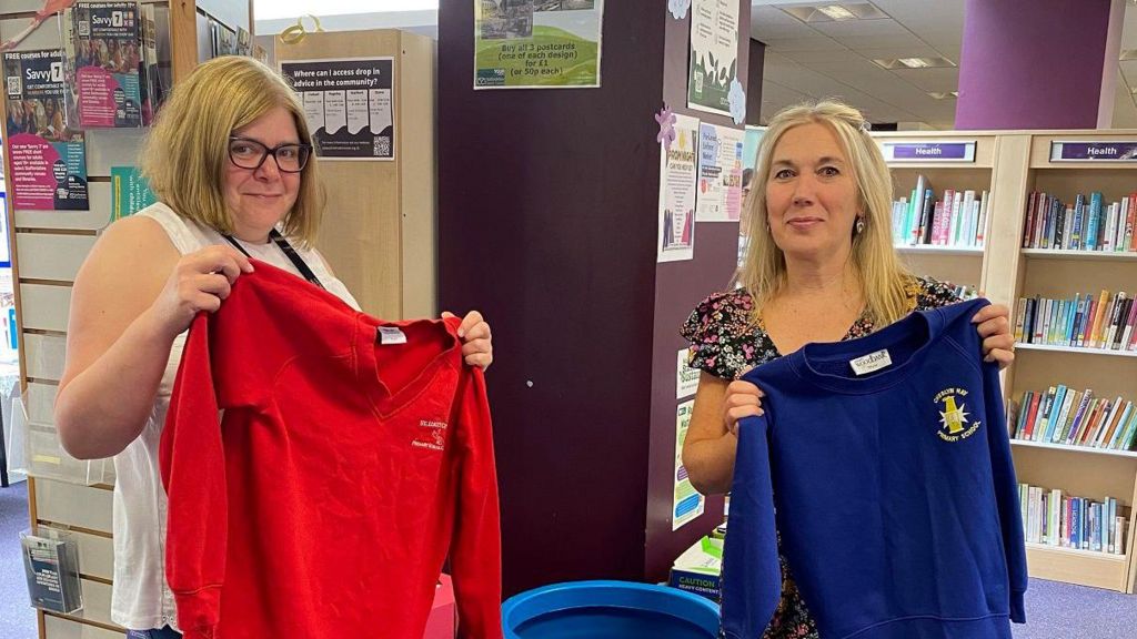 Two women stand in a library, one holding up a red school jumper and the other holding up a blue jumper