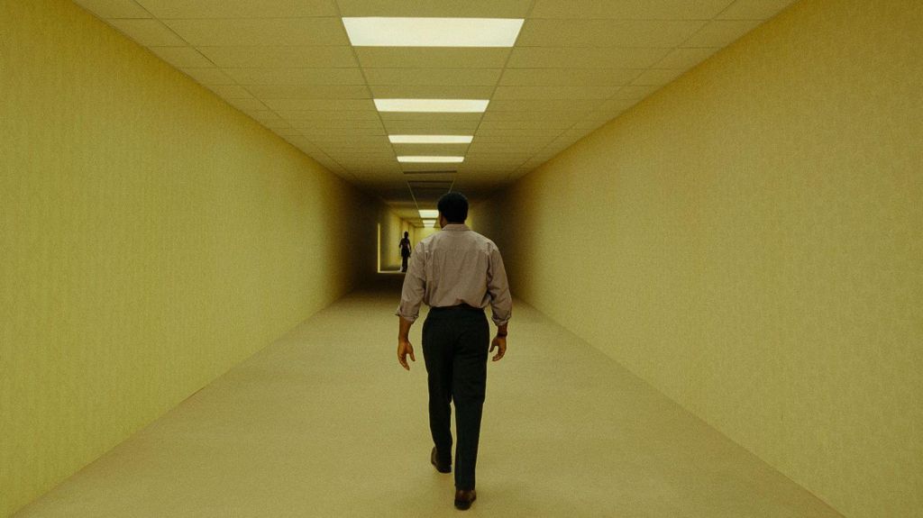 British actor Chiwetel Ejiofor as Clark, walking through the yellow hallways of the Backrooms, with a shadowy figure in the distance.