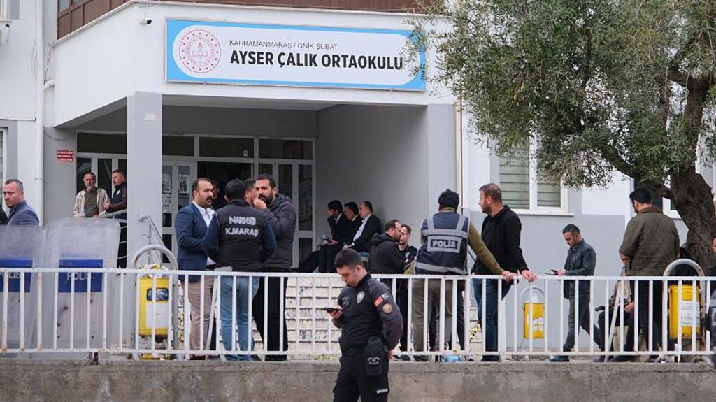 About two dozen men stand outside a school building, which has a sign reading 'Ayser Calik Ortaokulu', wearing a mix of plain civilian clothes and police uniforms.