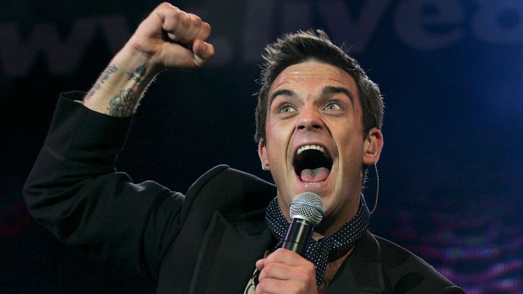 Robbie WIlliams with a microphone and his mouth open and his fist in the air at the age of 31, in 2005