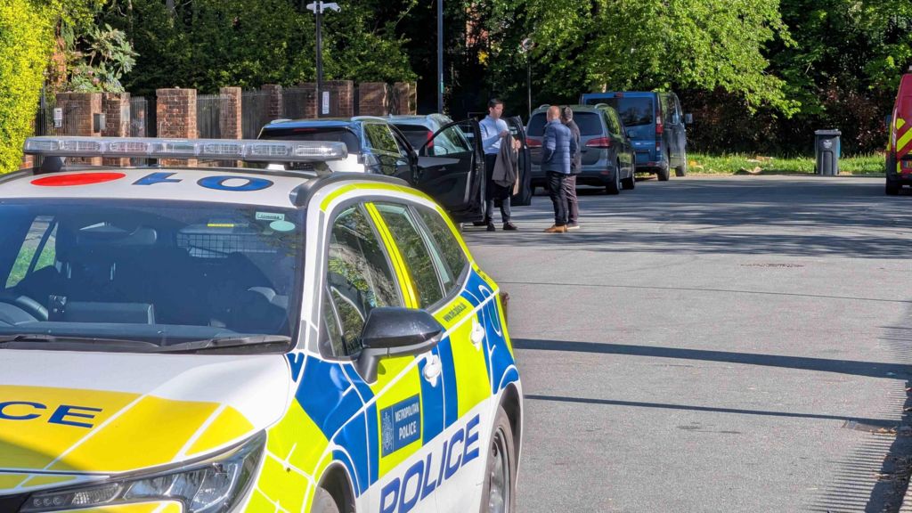 A marked Metropolitan Police car is parked on a residential street in north‑west London while emergency vehicles and several people stand beside parked cars further up the road, with trees and brick walls lining the street.