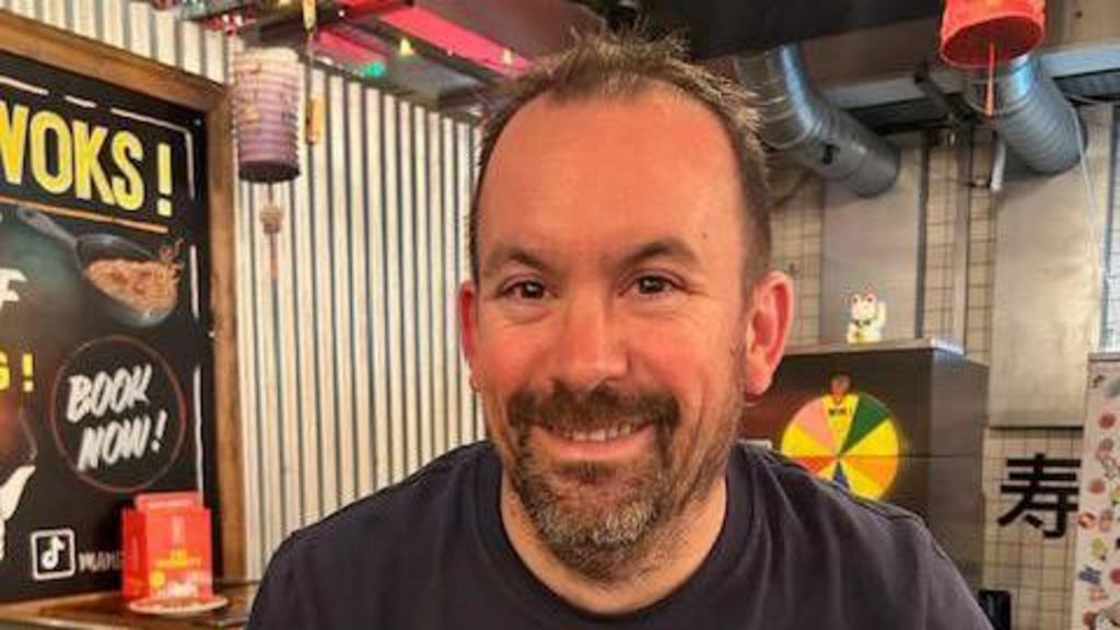 A man with short brown hair and a brown-grey beard sitting in a Chinese restaurant. He is smiling at the camera.