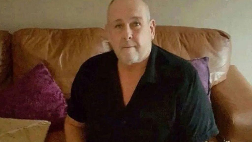 Steve Dymond looks directly at the camera, he is a sat on a brown leather sofa which has deep purple cushions on it. He wears a short sleeved black polo top