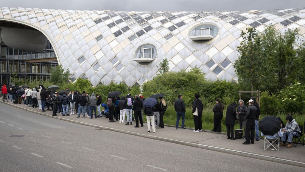 A line of people stand or sit on the pavement outside the headquarters, which has a shiny metal curved exterior.