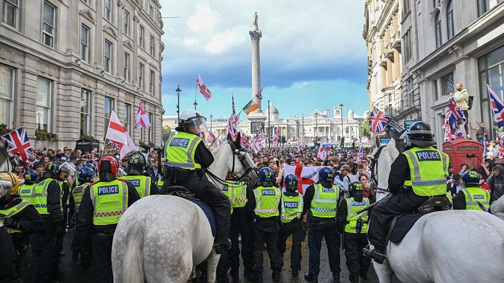 A row of police officers, including two on horseback, wear high vis jackets and riot gear in front of a large crowd of protesters near Nelson's Column.