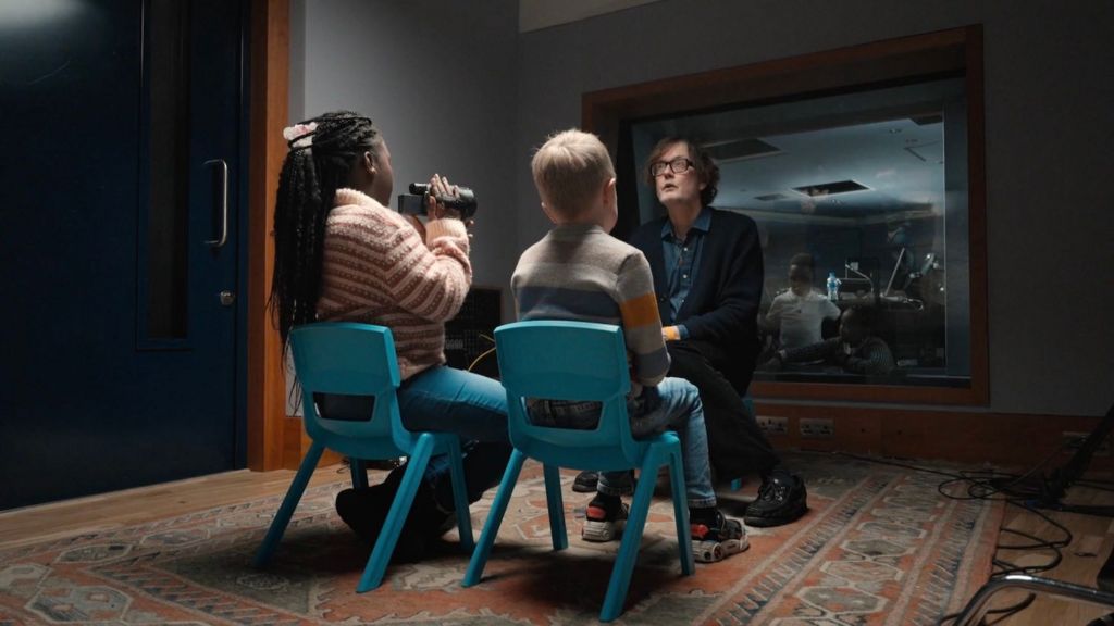 Jarvis Cocker is filmed and interviewed by two children sitting on blue plastic school chairs, in a studio at Abbey Road