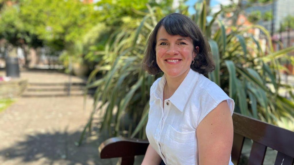 Labour candidate Rachel Blake, who has dark hair, is wearing a sleeveless white shirt and is sitting on a bench, smiling at the camera
