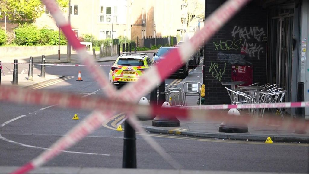 Red police tape crosses the road. Silver chairs are on the pavement opposite, inside the police cordon with a police car with fluorescent yellow marking parked beyond it.