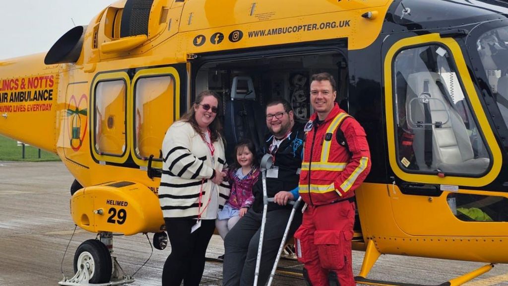 Four people stand in front of a yellow air ambulance helicopter. Dr Adam Chesters is wearing a red flight suit and is standing next to Tony who is dressed all in black and carrying crutches. Poppy, a little girl wearing a purple coat and blue dress smiles next to them. The group are joined by a woman with dark hair who is wearing a cream and blue striped jumper.