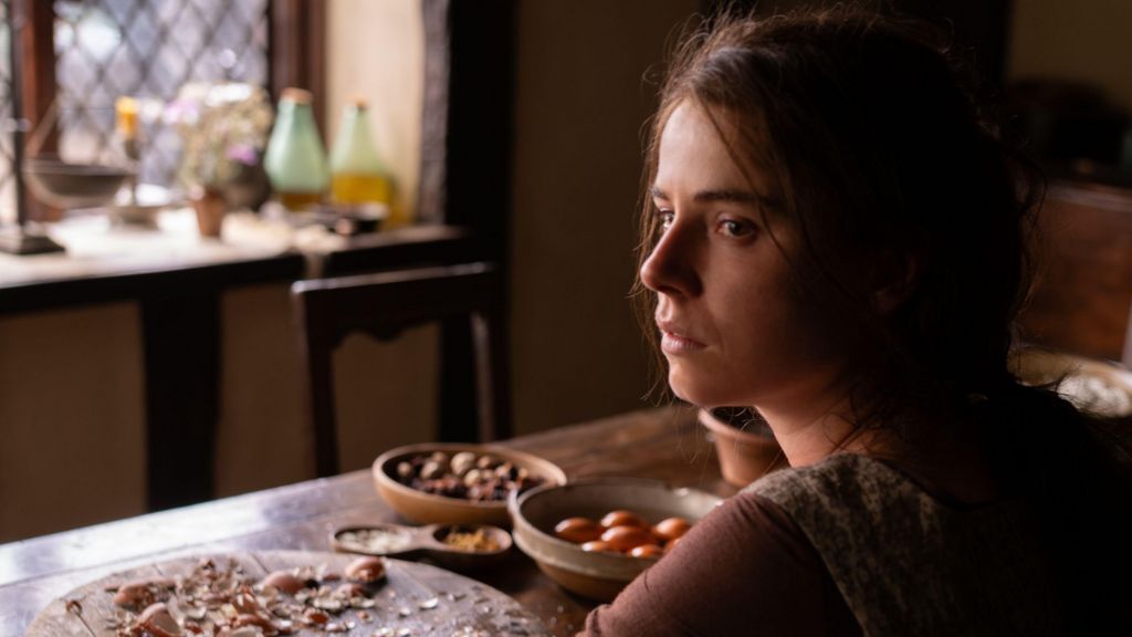 Jessie Buckley as Agnes in Hamnet, dark hair and a pensive expression, sitting at a table with food on it