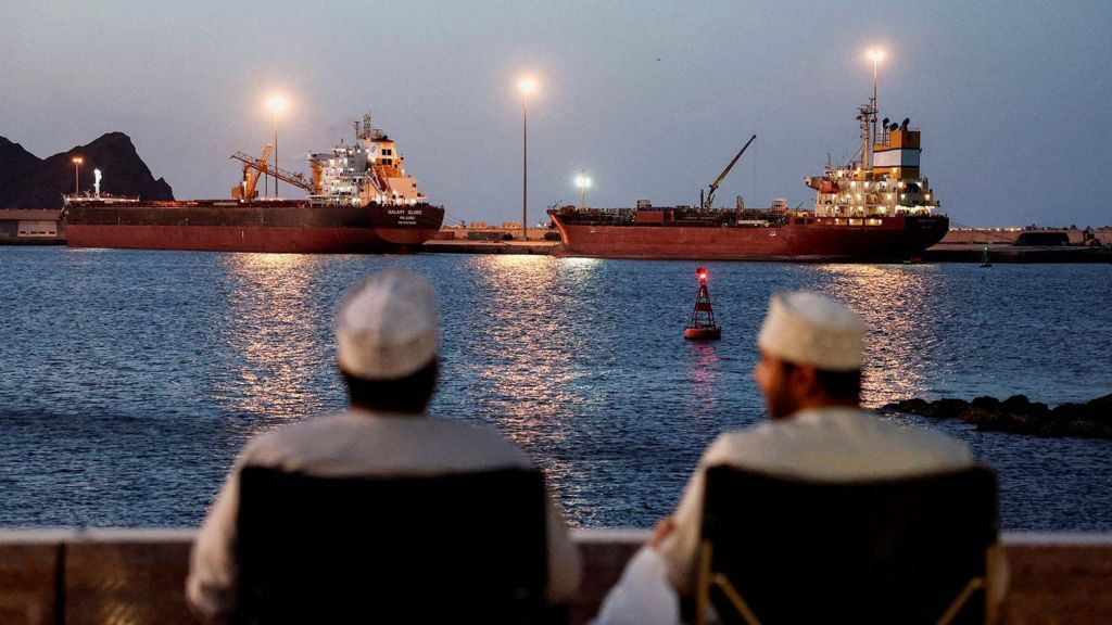 The backs of two men wearing white can be seen looking out to sea, where the Galaxy Globe bulk carrier and the Luojiashan tanker sit anchored as Iran vows to close the Strait of Hormuz, amid the U.S.-Israeli conflict with Iran, in Muscat, Oman, March 9, 2026. REUTERS/Benoit Tessier/File Photo
