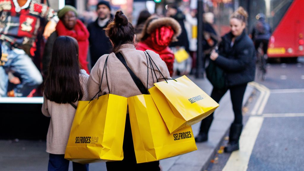 A woman walks along Oxford Street in central London with three bright yellow Selfridges shopping bags on her shoulders.