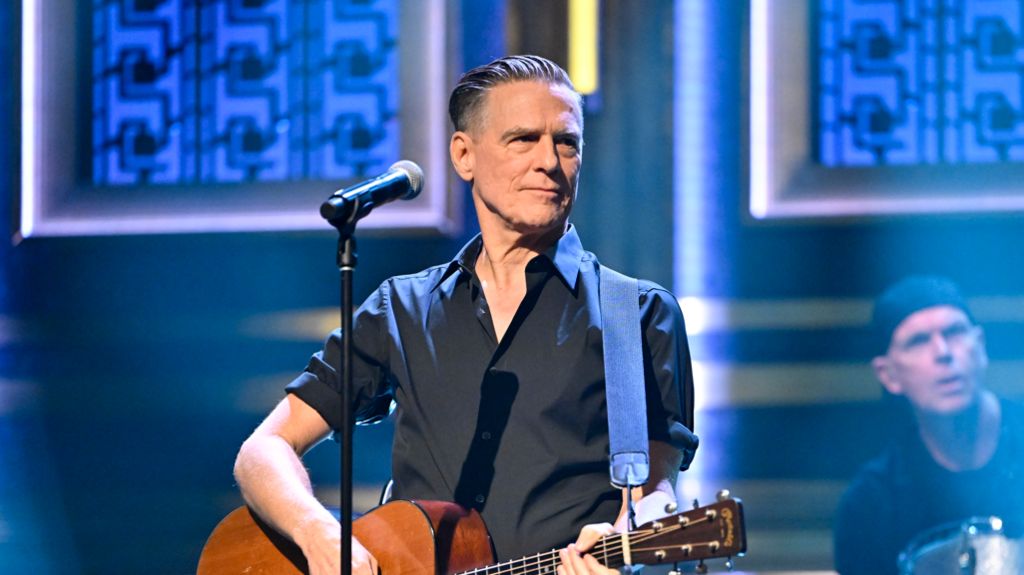 Bryan Adams stood in a black shirt playing a guitar, his drummer is behind to the left of him
