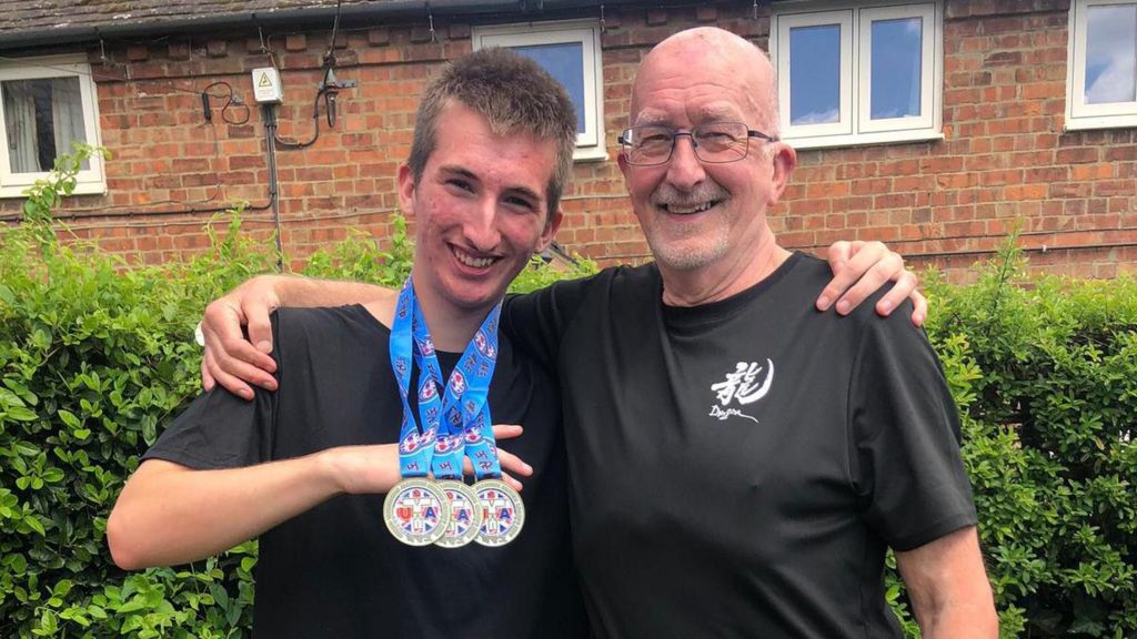 Dylan stands on the left, he is in a black t-shirt and is holding three gold medals, which are around his neck. They have blue ribbons with red, gold, white, and blue detailing. His dad, Tony, stands to his right. They have their arms around each others shoulders. Tony is wearing a black t-shire. He has a bald head and glasses.