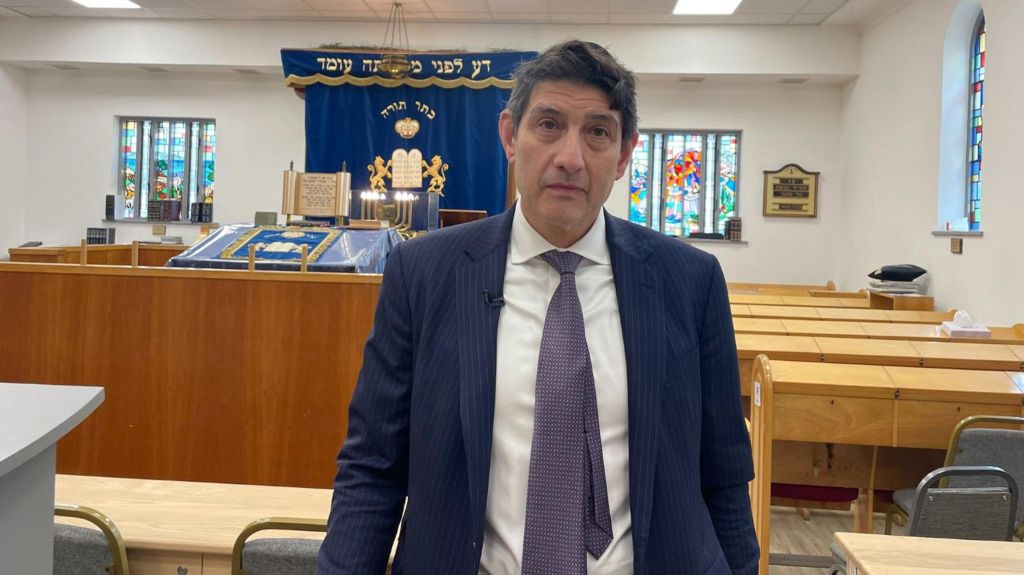 President of the Machzike Hadath synagogue Damon Hoff wears a navy suit, a white shirt and a purple tie as he looks directly towards the camera. Against the backdrop is the inside scene of a synagogue