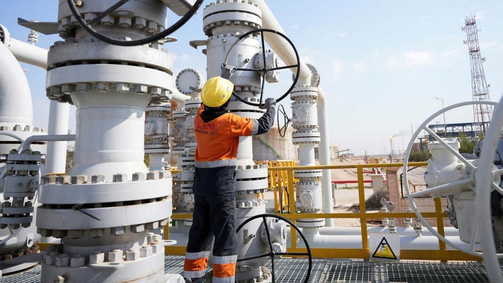 A worker operates valves at the Rumaila oil field. He is earing an organge boiler suit and yellow helmet . He is outside and there is a blue sky