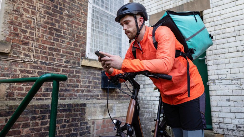 A delivery rider wearing an orange jacket rests his elbows on the handlebars of his electric bike while looking at his smartphone