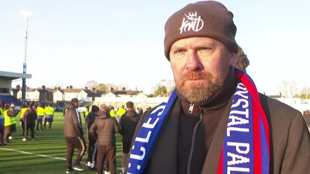 Anthony Dearnley is wearing a beige-coloured woollen hat and a commemorative match scarf and brown top and jacket is standing next to the pitch.