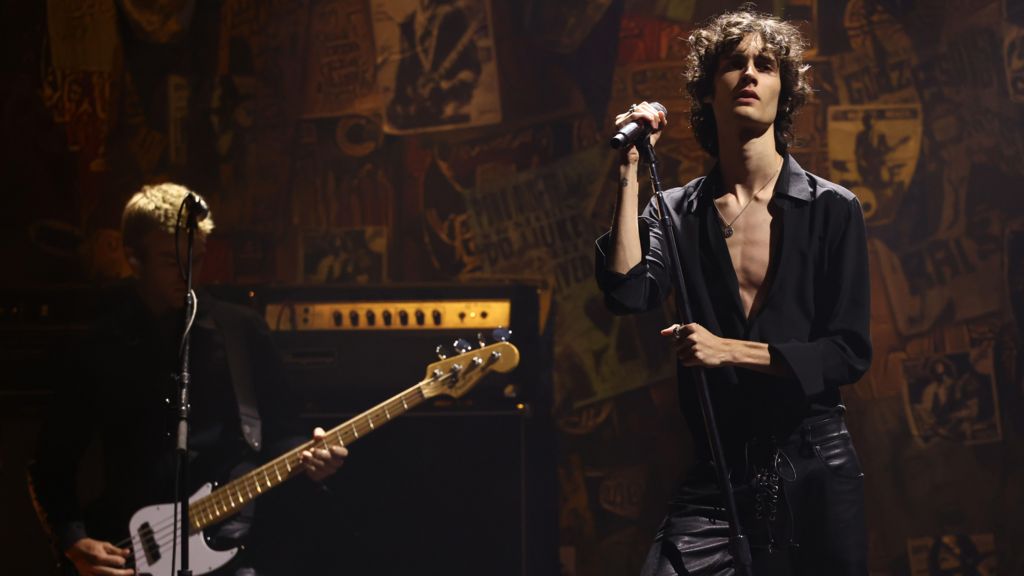 Sombr in a half-unbuttoned black shirt and black leather trousers holding a microphone stand and looking moody on stage next to a guitarist at the 2025 MTV Video Music Awards