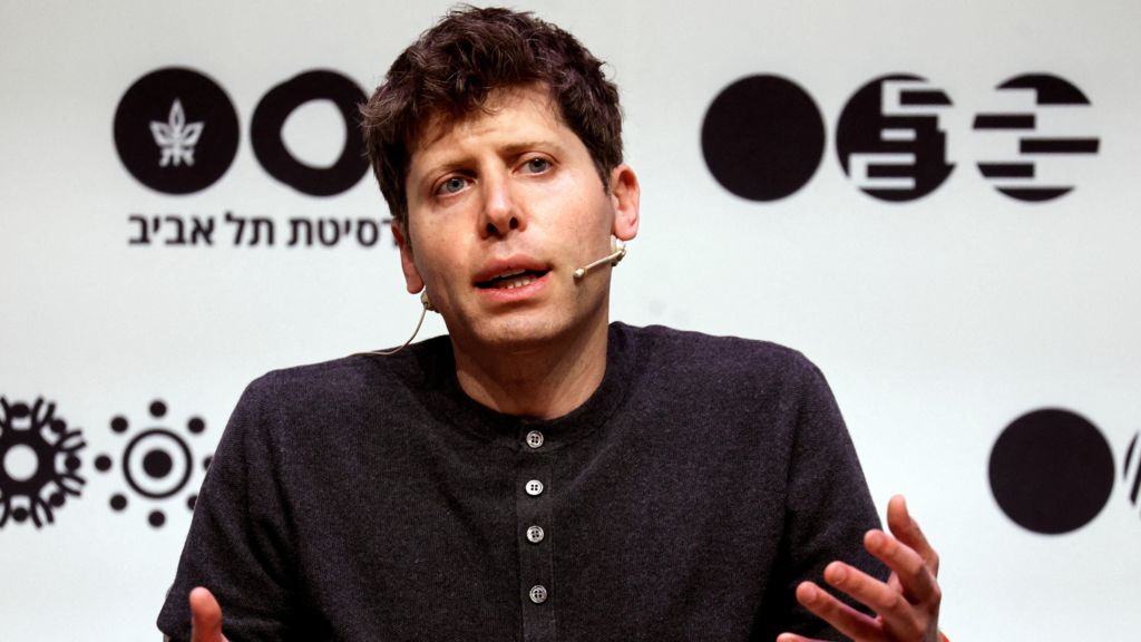 OpenAI leader Sam Altman sitting on a stage, speaking and gesturing with his hands while wearing a dark grey henley sweater.