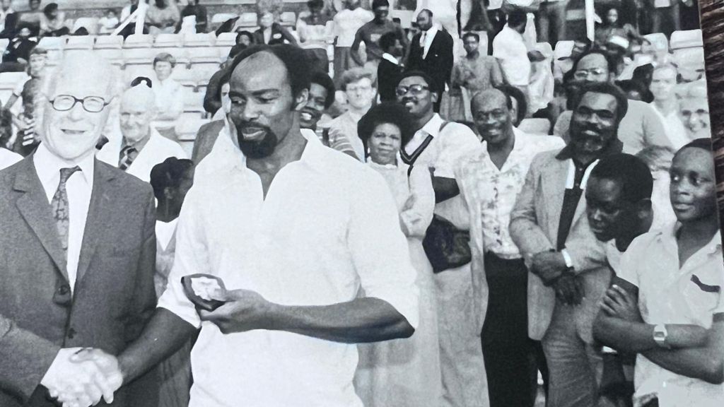 A black and white image from 1982, showing cricket coach and former Brixton West Indies player and captain Neville Walker shaking hands with a man, as he holds a plaque. The photograph is taken at a cricket ground, and spectators can be seen standing directly behind the pair, whilst some can be seen sitting in the background.”