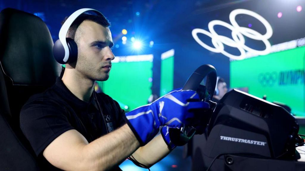 A young man wearing white over-ear headphones sits in front of a videogame controller steering wheel in an arena setting He wears an expression of concentration as two large screens glow green behind him and a large set of white Olympic rings lit in white hovers over the stage