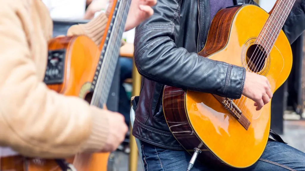 Two people are playing acoustic guitars outside. One is in focus and the other is blurry. One of the people is wearing a beige jacket and the other is wearing a leather jacket.