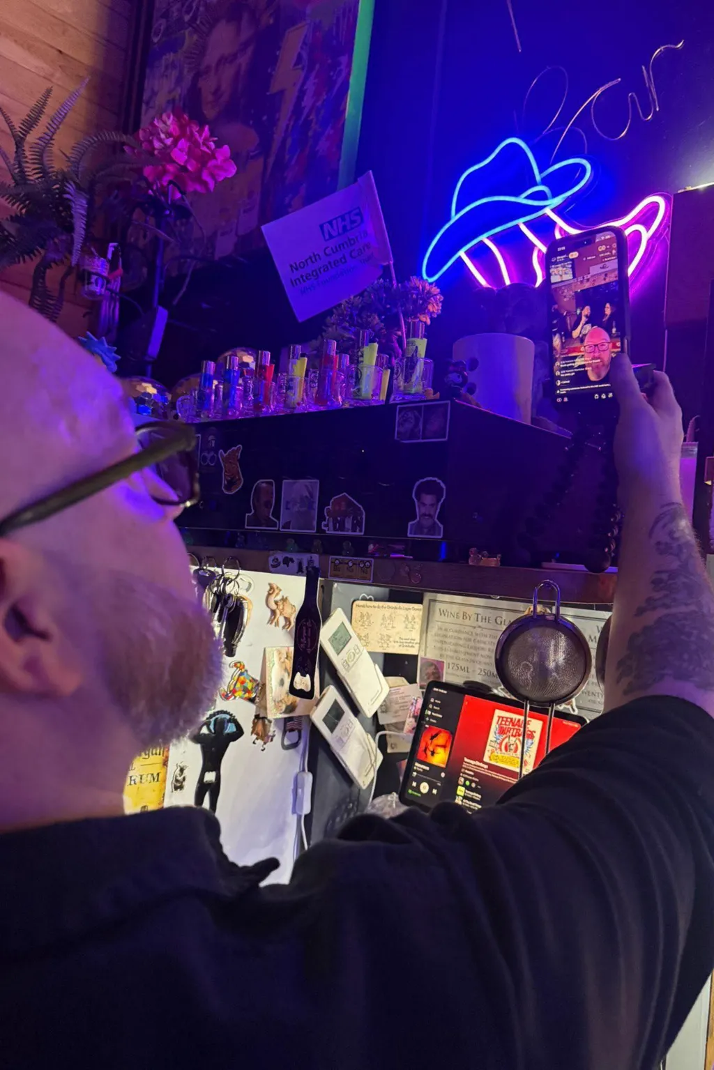 Morgan attending to a smartphone recording the livestream in his bar. The phone is held in a tripod and situated on a shelf next to bottles of vodka. A bright neon sign shaped like a cowboy hat glows blue and pink above the shelf.