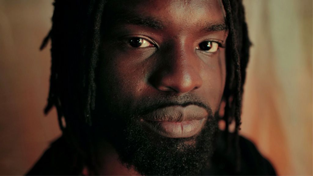 A close-up of Meji Alabi, with beard and dreads, looking into the camera