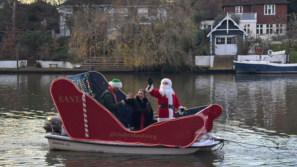 A sleigh mounted on a small motorboat carries people dressed as Father Christmas and two others along the River Thames, with houses and moored boats visible on the far bank.