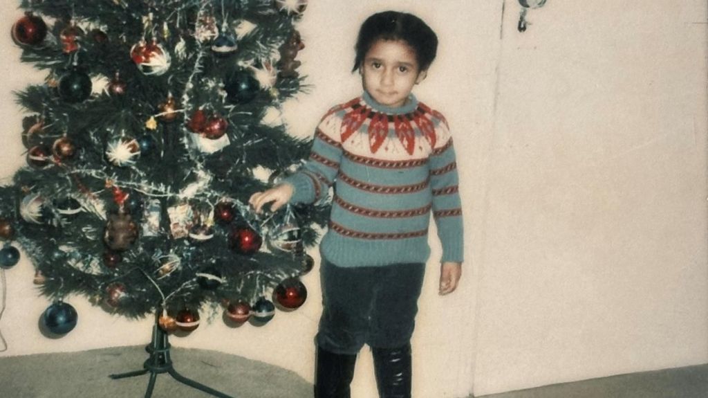 A polaroid image of a Andrea Bernard standing next to a decorated Christmas tree. She is wearing a patterned blue, white, and red knit jumper, dark trousers, and black boots.