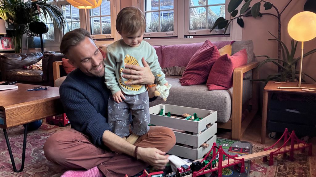 A father and son play with a wooden toy train set. The boy stands on the mans lap in a well decorated cosy living room. 