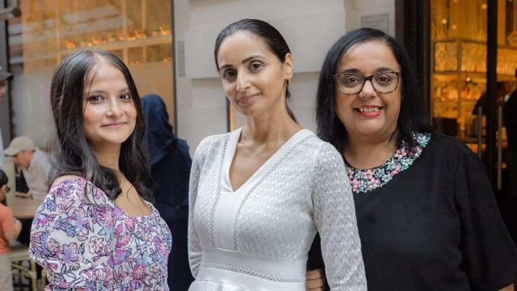 Three women smiling at the camera, the woman on the left is wearing a floral shirt, the women in the middle is wearing a white long-sleeve shirt and the woman on the right is wearing a black long-sleeve shirt with a floral collar.