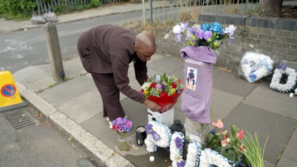   
Jodian places a bouquet of flowers at a street memorial to her son Daejaun, made up of floral tributes, candles, and a purple hoodie wrapped around a pole with a printed photo attached. The memorial sits along a pavement beside a brick wall and metal fence.