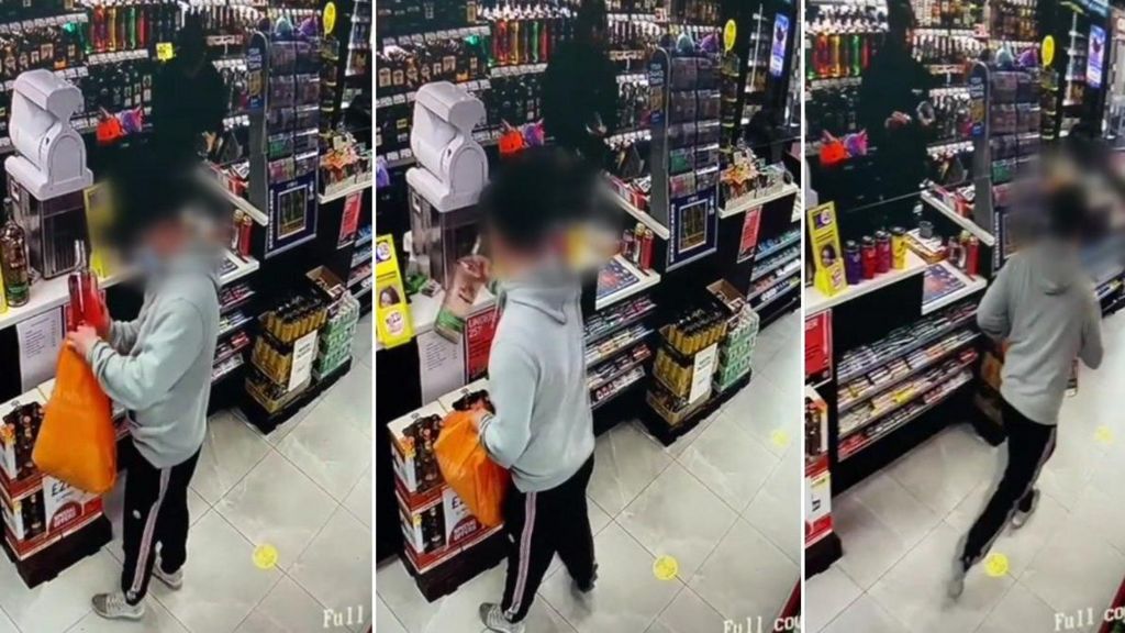A man, in a grey goodie and cap, puts two bottles of alcohol into an orange carrier bag and walks out of a shop