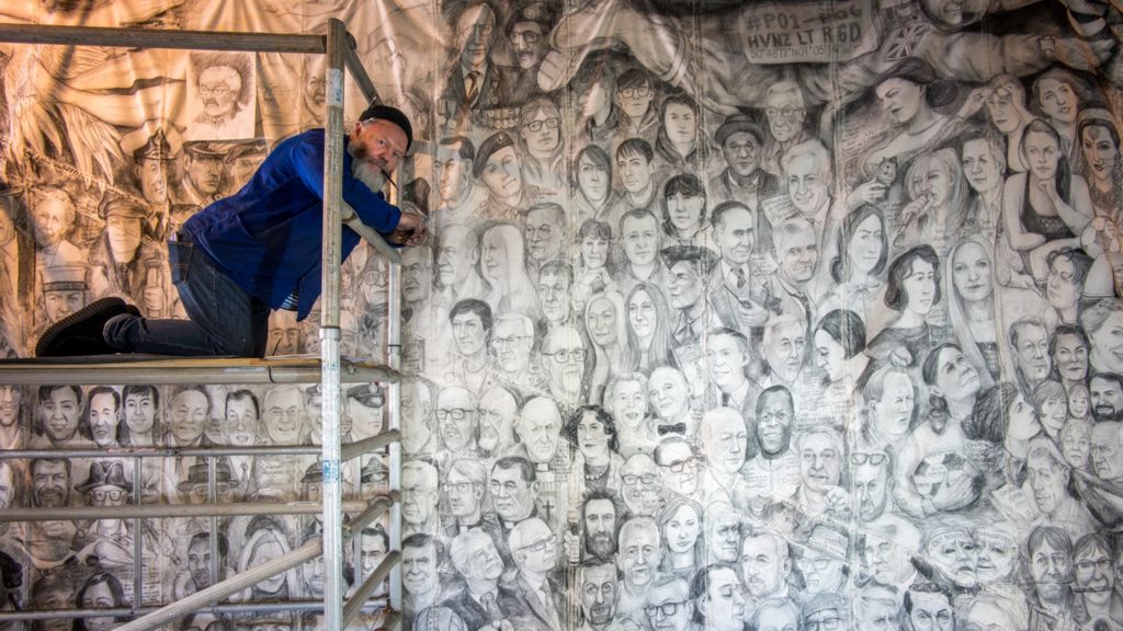 A man kneeling on scaffolding. He is sitting in front of a chalk mural which covers the whole wall. The artwork is made up of people's faces.