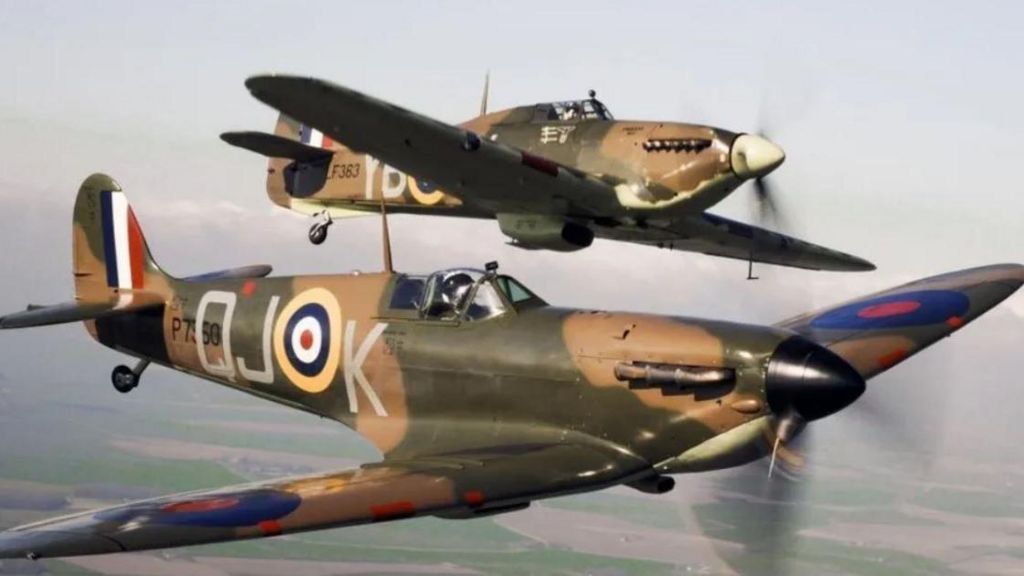 A Spitfire and Hurricane, both painted in camouflage colours, pictured flying together. The Spitfire is flying level, with the Hurricane banking slightly to port.