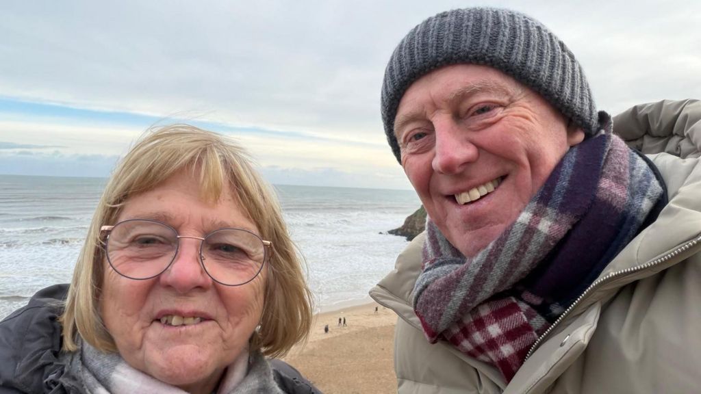 David and Barbara, a couple in their sixties, on a beach in winter. David is wearing a grey woolly hat and plaid scarf. Barbara has blonde hair and glasses. Both are wearing winter coats.