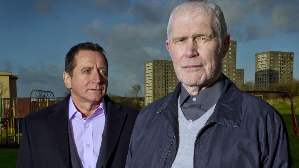 Alexander Morton on a blue jacket and jumper with a grey look serious while Frank Gallacher stares at the back of his head. There are a number of tower blocks behind them.