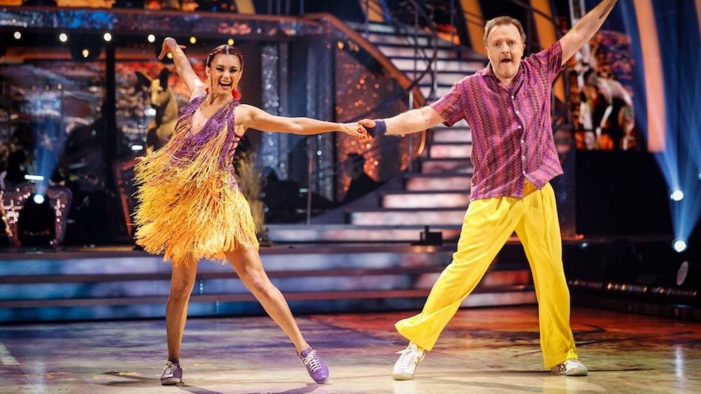 Dianne Buswell in a yellow dress with purple accents matches Chris McCausland in a purple shirt and yellow trousers.